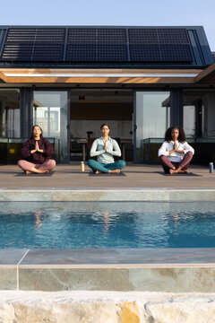 Three female friends sitting cross-legged on wooden poolside deck with yoga mats, reusable bottles