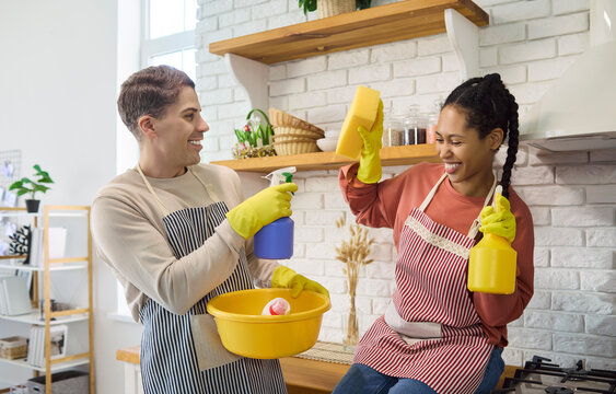 Smiling couple cleaning kitchen with gloves and aprons. They use spray and sponge with a bucket, making chores and housekeeping easy in a modern home. Teamwork and joyful home care concept.
