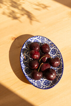 overhead shot of deep red cherries on a Portugese handmade blue and white dish on a light wood table with dappled shadows