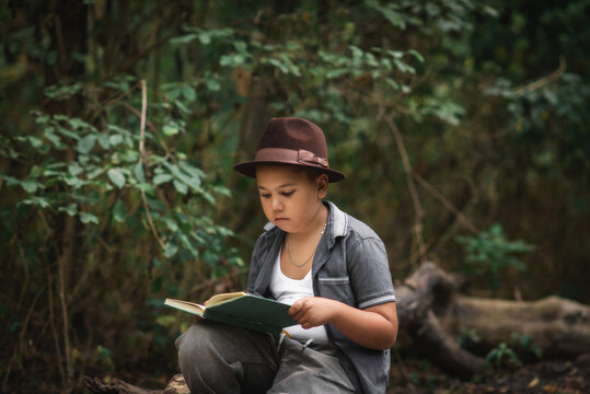 Boy reading an open book sitting in a forest. Thoughtful little boy wearing a hat sitting on the ground and reading an open book during summer in a forest.
