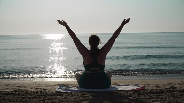 Silhouette of plus size woman sitting in lotus pose, raising arms and joining palms against sea. Morning yoga on sandy beach, wellness practice, flow of lifting arms up, joining palms, lowering.