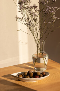 olives in a ceramic handmade plate of olives garnished with salt and dried herbs on a light wood table in afternoon light with strong shadows and a clear glass vase of minimal flowers