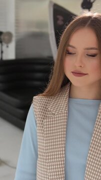 Vertical video. Attractive young model with long brown hair posing for the camera in a photo studio. The confident woman is wearing a stylish light blue top and a fashionable brown houndstooth vest