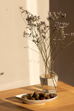 olives in a ceramic handmade plate of olives garnished with salt and dried herbs on a light wood table in afternoon light with strong shadows and a clear glass vase of minimal flowers