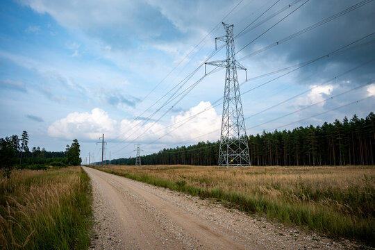 High voltage power transmission tower and overhead electricity lines beside a gravel road through a rural pine forest landscape