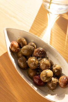 overhead shot of olives in a ceramic handmade plate of olives garnished with salt and dried herbs on a light wood table