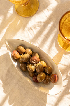 overhead shot of mustard gold wine glasses on linen tablecloth with ceramic handmade plate of olives garnished with salt and dried herbs with dappled shadows in afternoon light