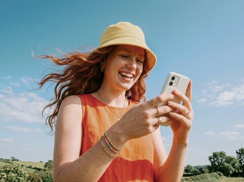Smiling woman with red hair laughing on phone in sunny field under blue sky