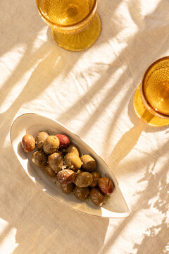 overhead shot of mustard gold wine glasses on linen tablecloth with ceramic handmade plate of olives garnished with salt and dried herbs with dappled shadows in afternoon light