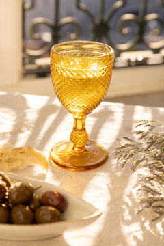 mustard gold decorated wine glasses on linen tablecloth with ceramic handmade plate of olives garnished with salt and dried herbs with dappled shadows in afternoon light and balcony in the background 
