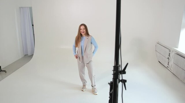 Beautiful young female model dancing and posing for the camera during a professional studio photoshoot on a cyclorama with bright lighting equipment, smiling and having fun