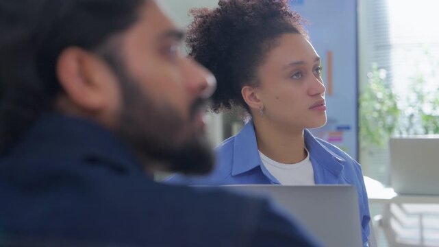 Thoughtful businesswoman listening intently during a meeting, seated behind a laptop with a colleague in the foreground, colorful data chart glowing softly on a screen behind her.