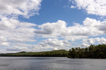 Fototapeta premium Scenic lake landscape with calm rippling water, lush green shoreline, resort buildings and gazebos surrounded by trees, with mountains visible in the distance under a dramatic cloudy sky. Peaceful out