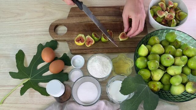 overhead view of a person cutting fresh green figs on a wooden board, baking ingredients including flour and eggs on a table, healthy homemade cooking process