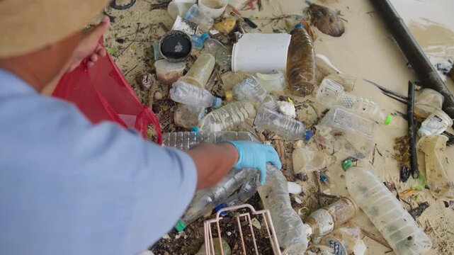 Man cleaning a sandy beach by collecting discarded plastic bottles and other trash, helping reduce pollution and promote environmental awareness, coastal conservation, and beach cleanliness.