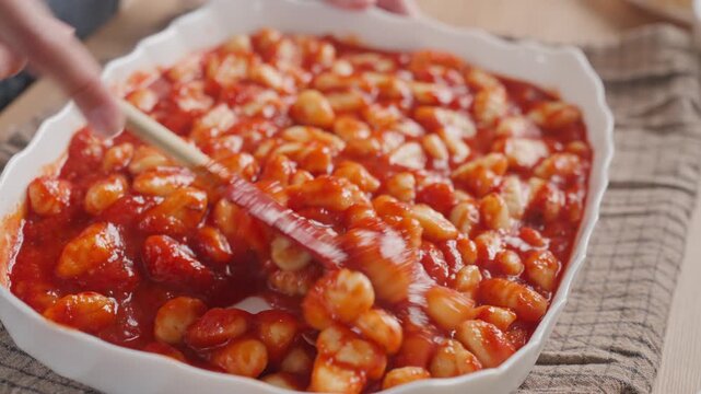 Woman stirs gnocchi in tomato sauce inside ceramic baking dish with spatula. Preparing homemade Italian style meal