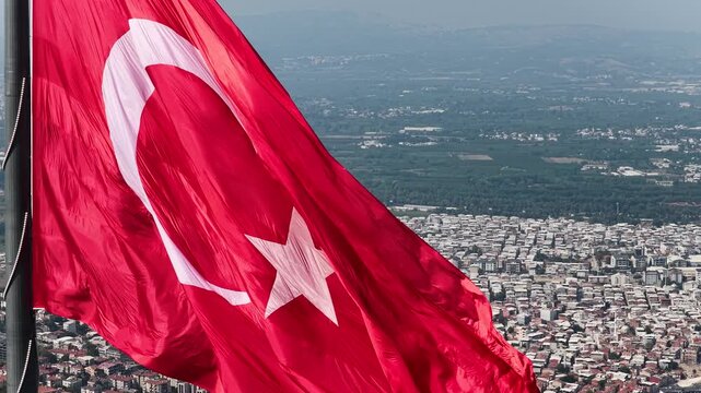 A close-up drone shot taken from the air of a massive Turkish flag proudly fluttering in the wind against a backdrop of city and plain scenery.