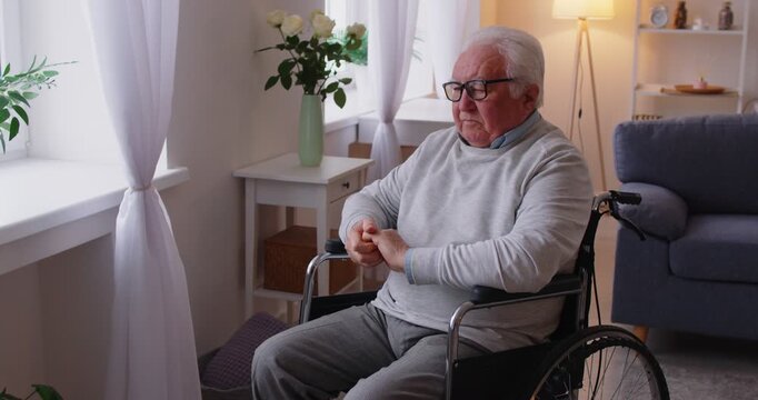Thoughtful Caucasian senior man, sitting in a wheelchair and looking out a window at home during rehabilitation. Introspection, recovery, and the challenges of living with a handicap.