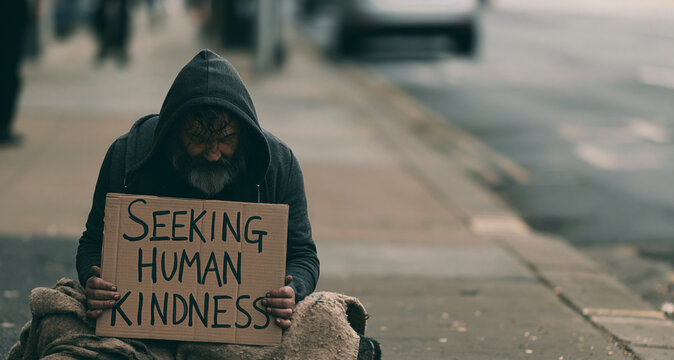  homeless man sitting on urban sidewalk holding a cardboard sign with text "Seeking Human Kindness", 
