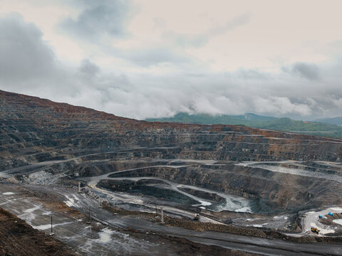 Open pit mine terraced excavation under cloudy mountain sky, mining industry aerial view
