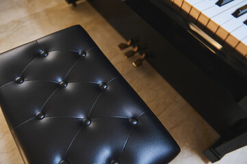 Black leather piano bench beside glossy grand piano with visible keys and pedals in a practice room © Taras Grebinets