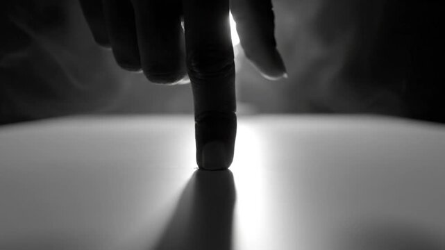 Dramatic black and white close up of a hand and finger touching a bright white surface with smoke in the background revealing a light bulb