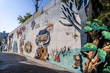 An artistic mural on a concrete wall in the fairytale village, depicting scenes from traditional Korean folktales with relief sculptures and colorful illustrations. © Gwibin