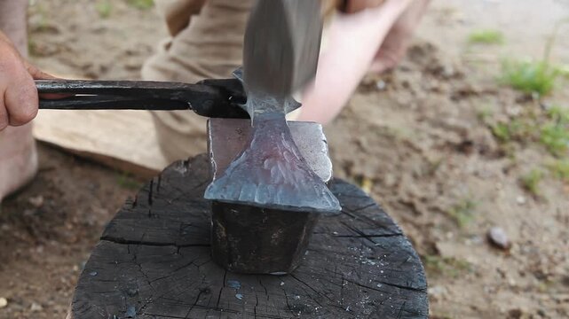 Blacksmith forging and shaping a traditional iron axe head with hammer on anvil outdoors