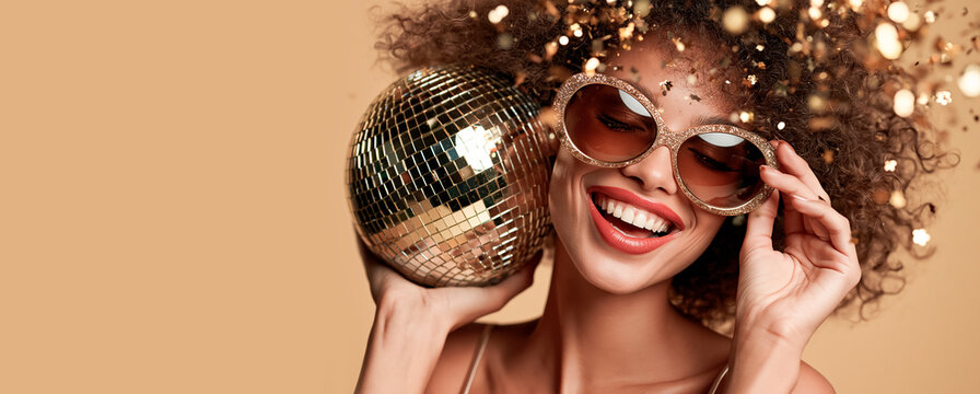 Happy beautiful positive young glamorous woman in glasses holding a disco ball, confetti flying in the background