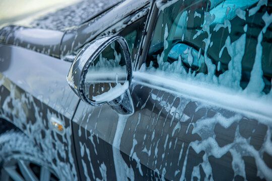 Thick detergent white soap foam dripping over a vehicle wing mirror during touchless washing at an automotive wash station