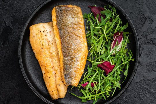 Pan fried sea bass fillets served on black plate with fresh arugula salad, gourmet seafood meal on black  background with copy space.