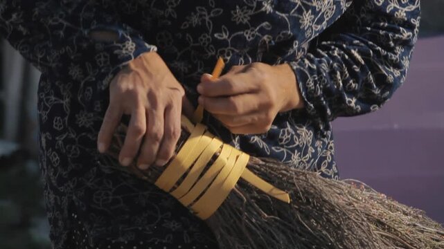 Hands weaving and cutting yellow plastic strip while making traditional handcrafted broom outdoors
