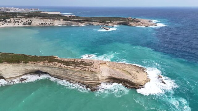 dramatic rocky headland breaking waves aerial drone footage malta offshore rock formation deep blue open sea distant town skyline turquoise coves mediterranean coastline scenic seascape