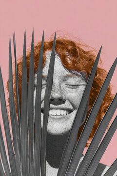 Freckled red haired woman smiling behind the spiked fronds of a monochrome palm leaf on a soft pink background