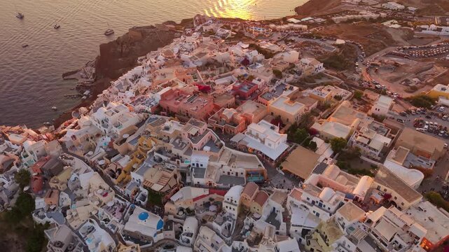 High angle drone footage looking down at Oia village street layout white rooftops and hillside architecture at sunset Santorini Greece