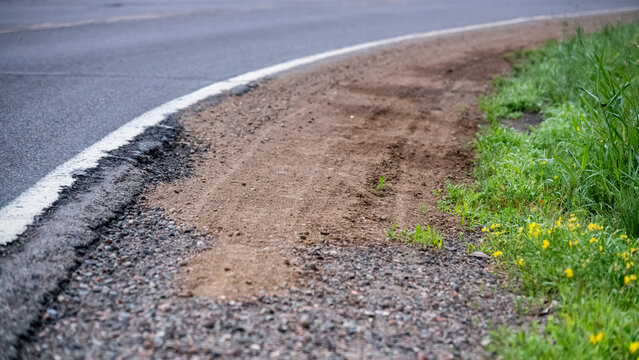 an unpaved, eroded dirt and gravel road shoulder adjacent to the edge of an asphalt highway, featuring tire ruts and edge drop-off.