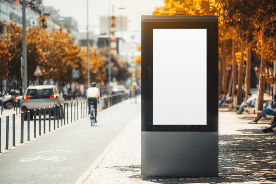 Blank vertical advertising display stands beside a sunny city bike lane, with a distant cyclist silhouette on the left among traffic, trees and warm urban background blur