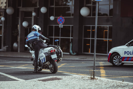 Rear view of an unrecognizable helmeted traffic police officer mounting a motorcycle beside a patrol car on a European city street, capturing an active law enforcement moment