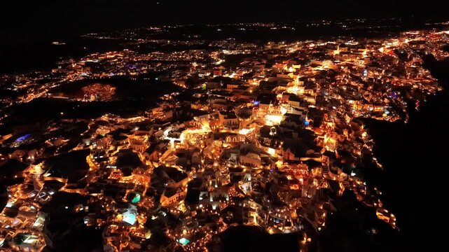 Aerial night drone footage of Oia peninsula glowing with golden city lights on dark cliffside in Santorini Greece