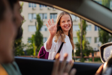 Naklejka na ściany i meble Cheerful girl waving to mother and father sitting inside car during morning school drop-off, viewed through vehicle window