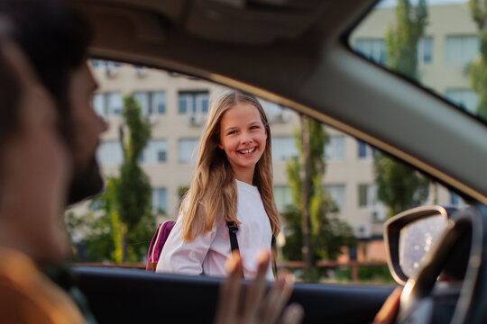 Happy schoolgirl smiling at her parents while they waving bye from inside vehicle after bringing her to school