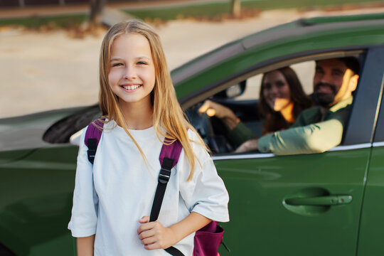 Happy schoolgirl with backpack standing in front of family car and smiling at camera while her parents bring her to school on sunny morning