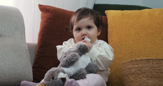 Cute baby girl sitting on couch smiling joyfully then playfully biting her stuffed bunny toy