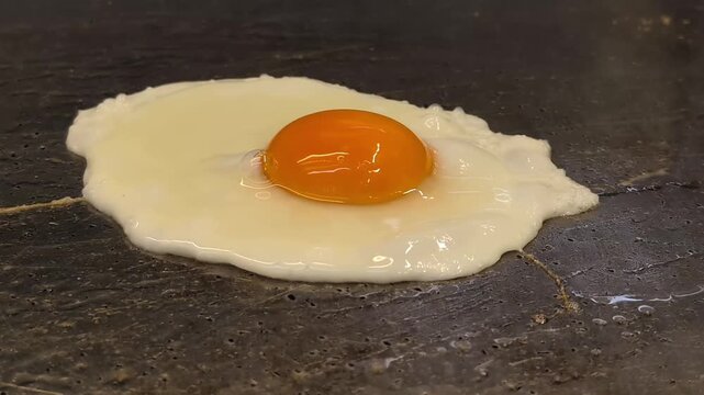 Close-up shot of a sunny side up fried egg with a bright orange yolk cooking on a hot flat top griddle. The egg white sizzles and sets while the golden yolk remains glossy and intact, capturing the cl
