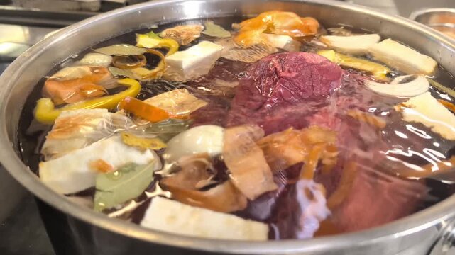 Close-up of a chef's gloved hand placing raw beef brisket into a large stainless steel pot filled with water, charred onion halves, carrots, celery root, leek, bay leaves and peppercorns. Traditional 