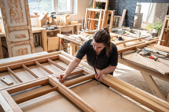 Female craftswoman emerged in work on restoring antique window frame at workbench in workshop, talented woman carpenter measures wooden frame parts and making marks on wood planks with pencil.