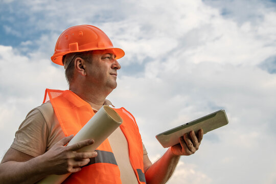 A construction worker stands outside with plans rolled up in one hand and a tablet in the other. He wears a safety vest and helmet. The clouds fill the sky in the background
