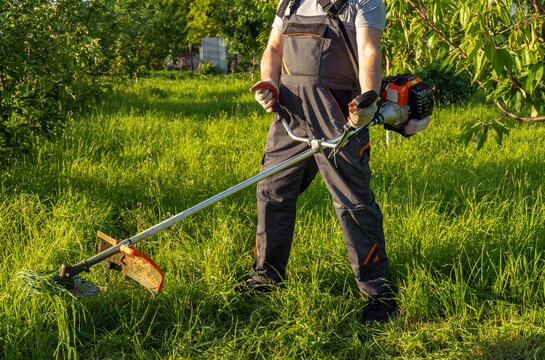 A worker stands in a green field holding a string trimmer. Tall grass surrounds him as he prepares to cut the grass on a sunny day in a rural area