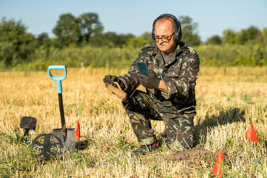 A man in military attire kneels in a field with a metal detector. He holds a phone in one hand and examines a dirt clump in front of him. Red flags mark his search area