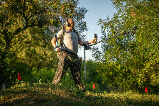 A treasure hunter stands on a small hill in a park using a metal detector. The sun is shining, and he is checking the ground while wearing headphones. 